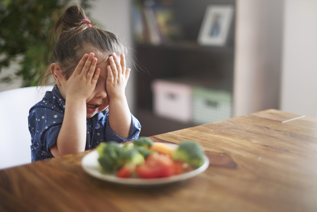 Child covering their face during a mealtime with vegetables on the plate, showing feeding reluctance.