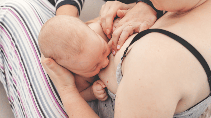 Infant breastfeeding with parent and clinician providing gentle feeding support, demonstrating safe latch and positioning