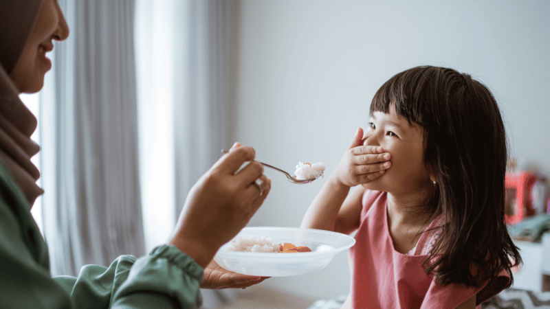 Child laughing while an adult offers food, representing playful mealtime interaction and positive feeding experiences.