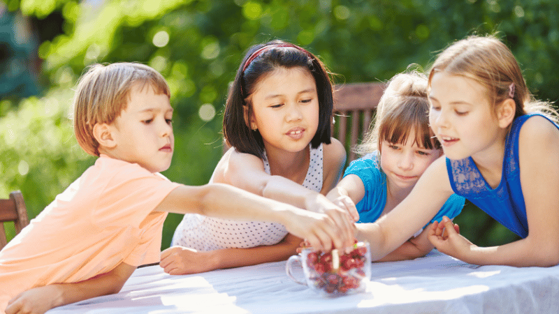 Children reaching into a container of cherries outdoors, representing food curiosity and sensory exploration.