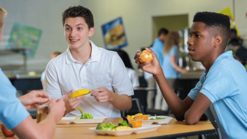 Teenagers sitting at a school lunch table eating fruit and vegetables, representing mealtime participation and social eating.