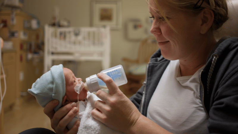 Adult feeding a newborn with a bottle in a hospital setting, representing early infant feeding support for babies needing medical care.