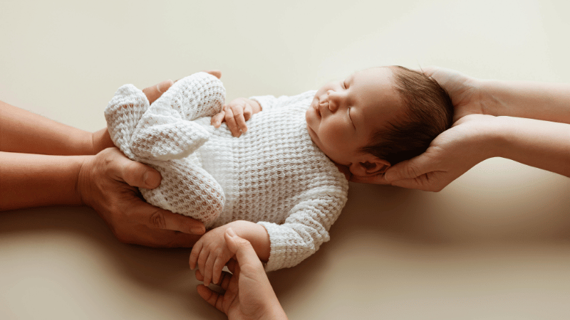 Newborn being gently supported by several caring hands, symbolising Thrive’s collaborative, family‑centred infant feeding approach.