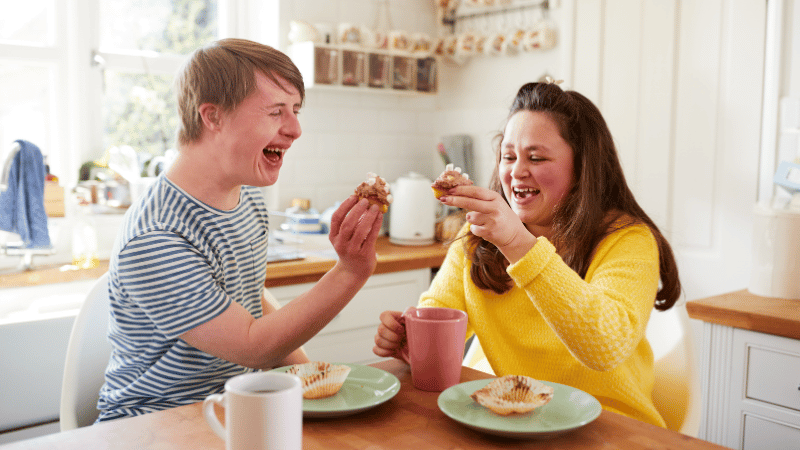 Two adults sharing cupcakes at a kitchen table, representing social eating and mealtime participation for NDIS participants.