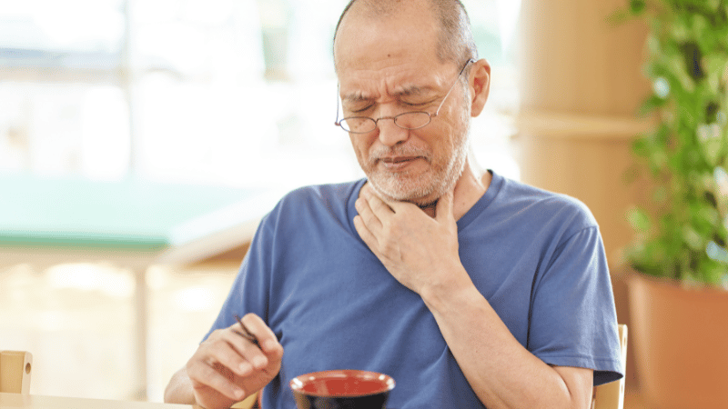 Older adult touching their throat while sitting at a table, representing swallowing concerns and dysphagia support for NDIS participants.