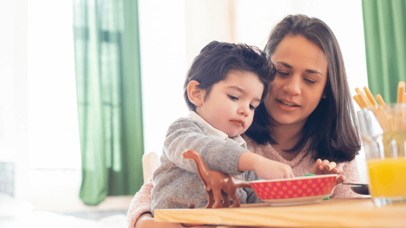 Toddler exploring food at the table with a toy nearby, representing early signs of feeding challenges in a safe, neutral way.