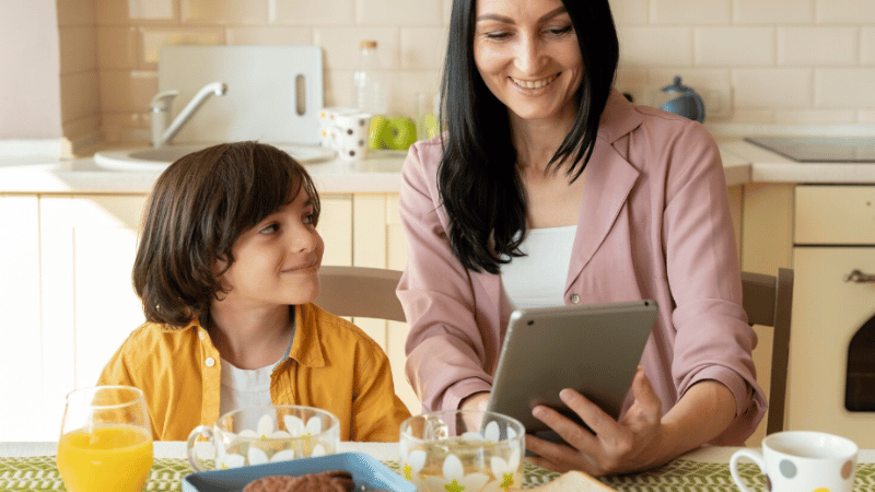 Adult and child sitting together at a kitchen table using a tablet, representing telehealth parent coaching for children and teens’ feeding support.
