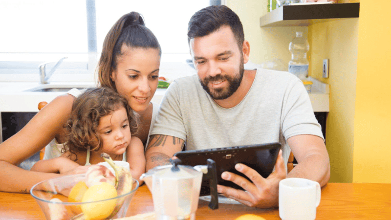 Parents and child sitting together looking at a tablet, representing telehealth parent coaching for toddler feeding support.