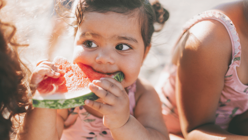 Toddler holding and eating a slice of watermelon during a relaxed, natural mealtime moment.