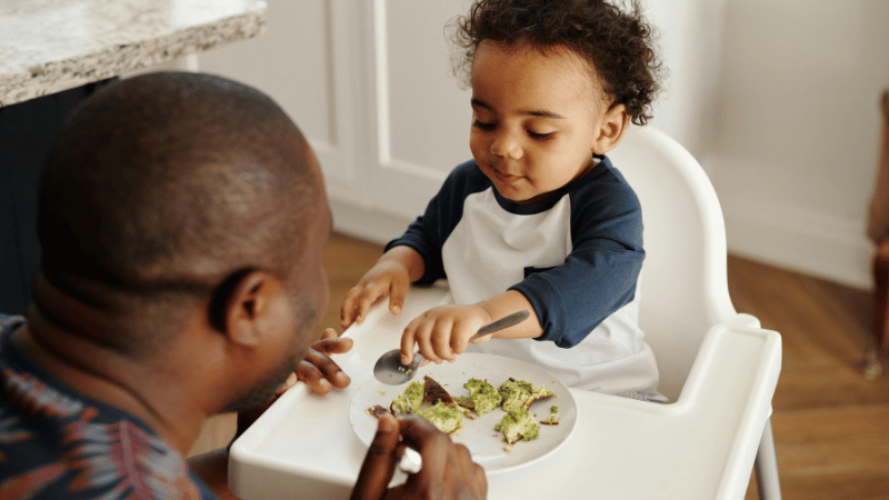 Toddler sitting in a high chair eating soft food with an adult nearby, representing support for selective eating, sensory feeding differences and mealtime participation.