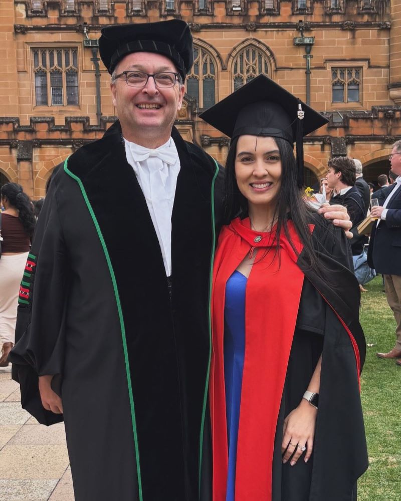 Dr Stefani Knijnik standing with a senior academic official in academic regalia outside a historic sandstone university building on her PhD graduation day.