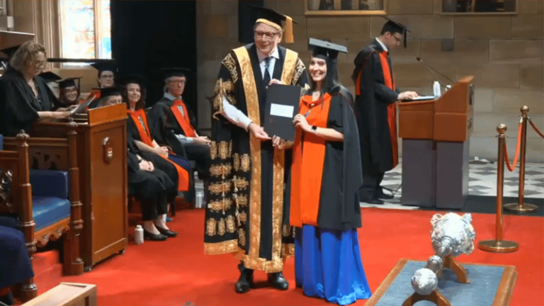A university graduation ceremony where a graduate in cap and gown receives her PhD certificate from an academic official in formal regalia.